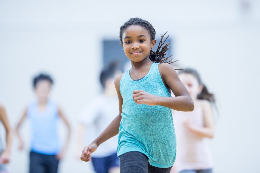 An african american girl runs and smiles at the forefront of a group of kids in her elementary physical education class.